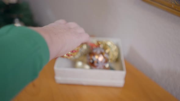 Female Hand Picks Up Glittery Christmas Bauble From A Box And Hangs It On A Christmas Tree alt