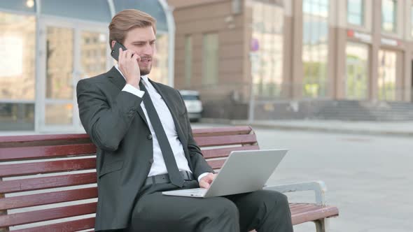 Businessman Talking on Phone and Using Laptop While Sitting Outdoor on Bench alt