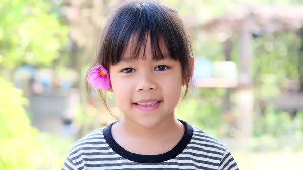 Cute little Asian girl with pink flower on ear. Childhood happiness concept. alt