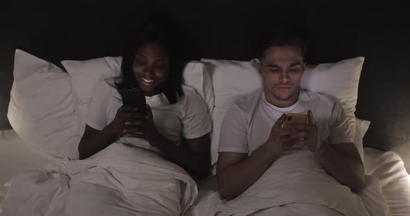 Happy Multi-ethnic Couple with Smartphones in Bed. People, Technology, Internet and Communication alt