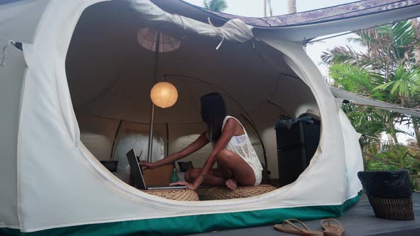 Pretty Woman Sitting Inside Luxury Tent House and Working on Laptop and Phone alt