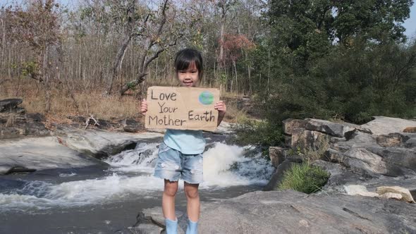 Portrait of cute little girl standing with Love Your Mother Earth poster at a waterfall in the fores alt