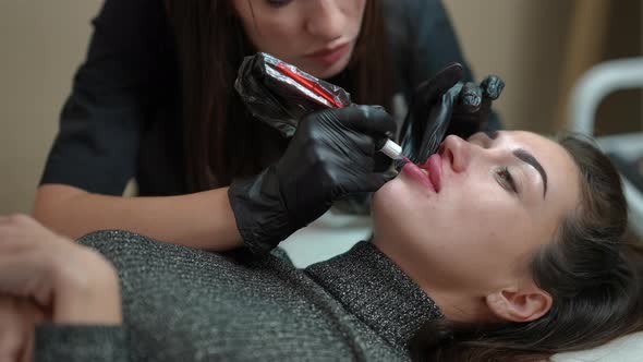 Portrait of Young Woman in Beauty Salon with Professional Tattoo Master Filling Lip Skin with Red alt