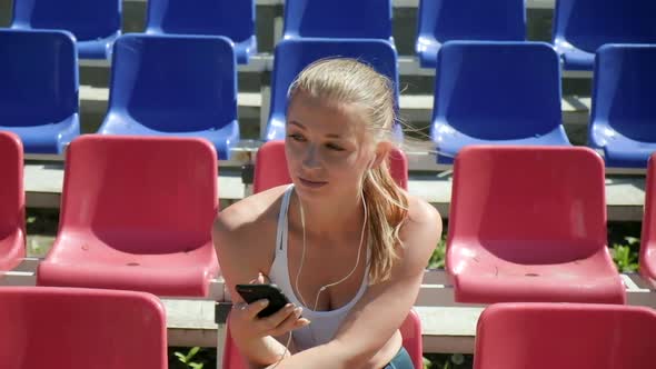 Young Caucasian Female with Headphones Listens to Music on Smartphone at Stadium