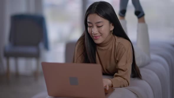 Smiling Asian Young Woman Lying on Couch Typing on Laptop Keyboard alt