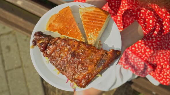 Young woman enjoying the view of grilled ribs on disposable plate. A slender athletic girl holds alt