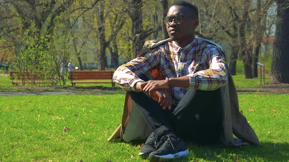 A Young Black Man in Glasses Sits on Grass in a Park on a Sunny Day and Looks Around alt