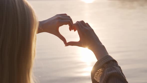Cute Blonde Woman Shows a Heart with Her Hands at Sunset By the Lake