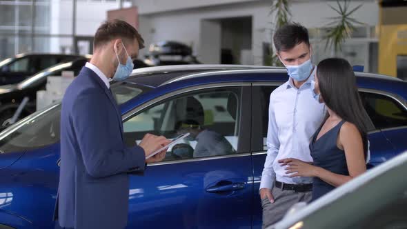 Young Couple in Masks Choosing Car at Dealership alt