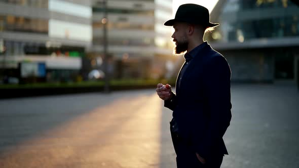 a Bearded and Mustachioed Man in a Hat Stands with a Cigarette in Hand on a Blurred City Street alt