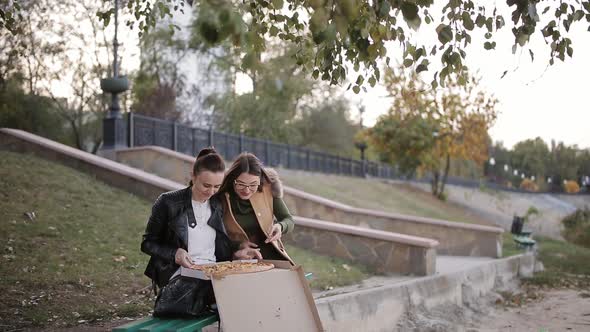 Two Women Eating Pizza in a Large Box in the City Sitting on a Bench on the Promenade