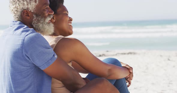 Happy african american couple sitting and embracing on sunny beach alt