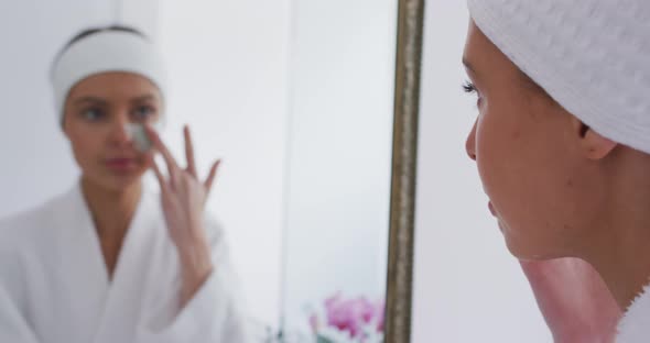 Woman in bathrobe applying face mask while looking in the mirror  alt