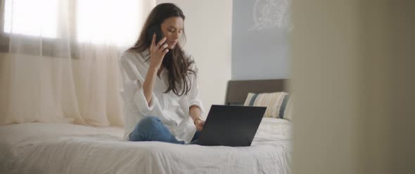 A woman talking to someone on the phone while working with a laptop from home.  alt