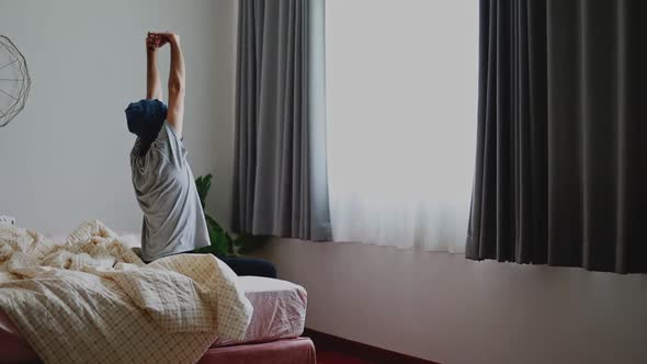 Attractive Asian man waking up and stretches while sitting on the bed in bedroom morning.