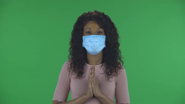 Portrait of Beautiful African American Young Woman in Medical Mask Is Looking Straight and Prays alt