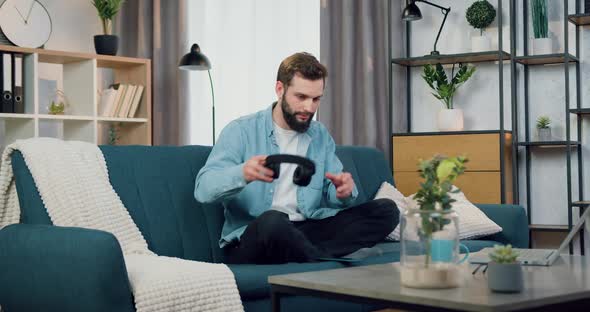 Man in Headphones Relaxing Under Favourite Music Sitting in Lotus Pose on Soft Couch at Home alt