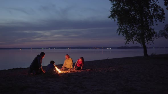 People Sit By Fire During Family Hike at Beach By Sea