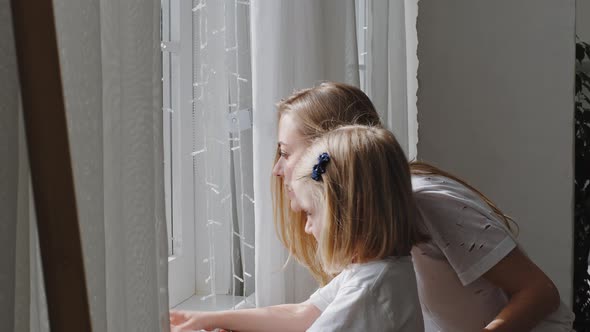 Young Caucasian Mother and Little Cute Daughter Child Girl Stand By Window Near White Curtains and alt