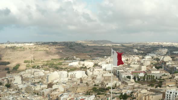 Flag of Malta Waving in Wind with View of City on Gozo Island, Aerial Close Up Slide Left alt