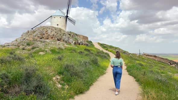 Young blonde country girl walking towards an old windmill in a meadow in sunny day alt