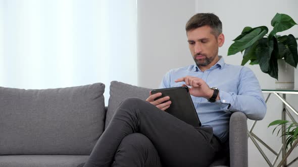 Businessman in Light Blue Shirt Sitting on the Couch Using Tablet Taping Screen alt