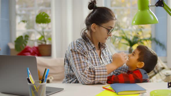 Young Mother Sitting at Desk with Laptop and Checking Temperature of Cute Little Son Feeling Unwell alt