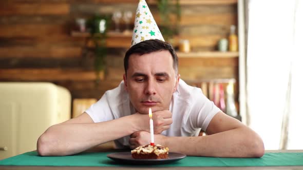 Lonely Man Sitting in Front of Little Cake with Lighted Candle and Blowing It. alt