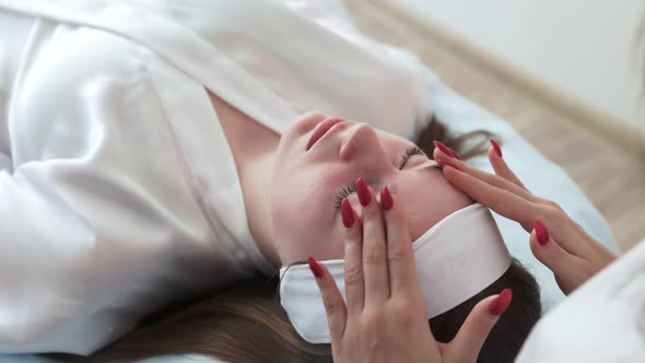 Hands of Beautician Making Massage on Face of Woman Lying with Closed Eyes on Couch alt