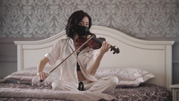 Beautiful Girl in Mask Playing the Violin While Sitting on the Bed at Home