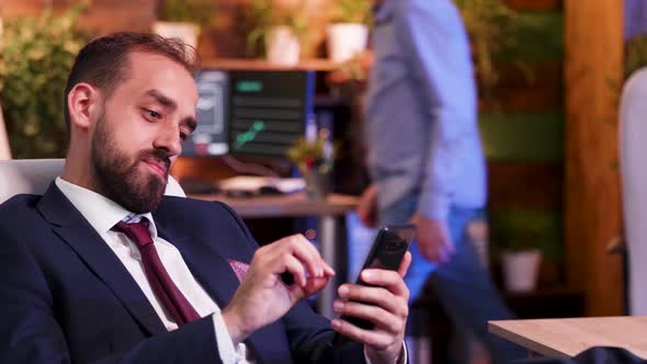 Relaxed Businessman in the Office at Night Time Scroll Down on His Phone alt