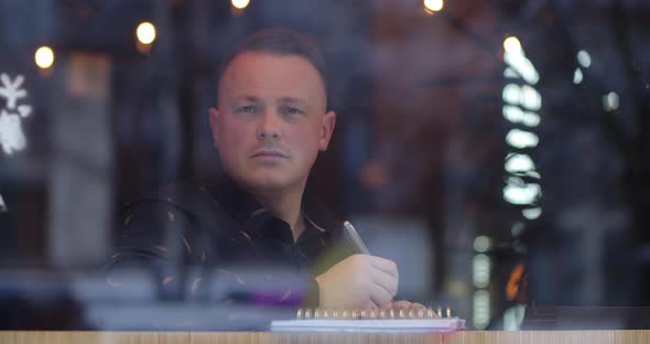 A Man Sits In A Cozy Cafe, Thinking About The Text And Writes It In A Notebook