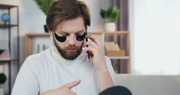 Young Bearder Which Touching Black Collagen Patches Under Eyes while Talking on Mobile alt