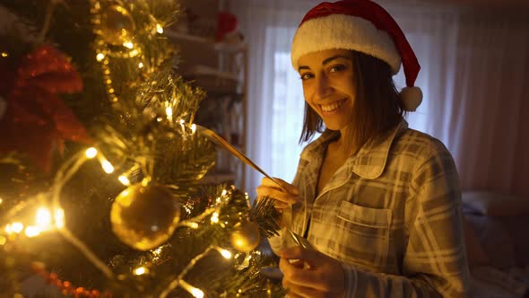 Slow Motion Portrait p of Happy Woman in Santa Hat Hangs Up Golden Ball on Branch of Christmas Tree alt