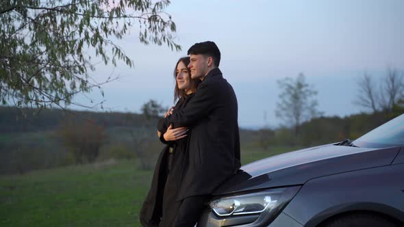 Happy Loving Couple of Travelers Stand Next to Car and Admire Beautiful Sunset alt