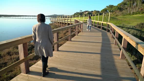 Two Females Walking on a Boardwalk Near a River Shore with People Ahead