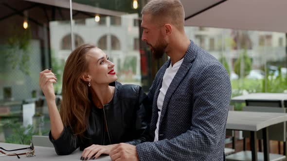 a Man with a Beard Looks at a Blonde with Bright Lipstick Then Takes Her Hand at a Cafe Table on the alt