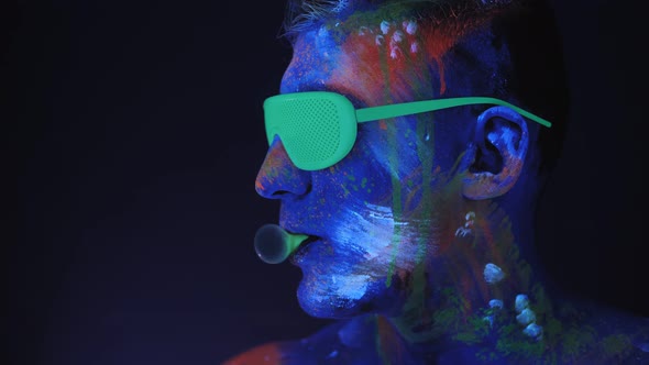 A Man with UV Drawings and Green Neon Glasses Inflates a Bubble of Bubblegum alt