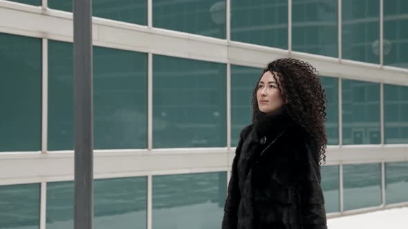 A Curlyhaired Stylish Middleaged Brunette Woman in a Black Fur Coat Walks Alone in the City on a alt