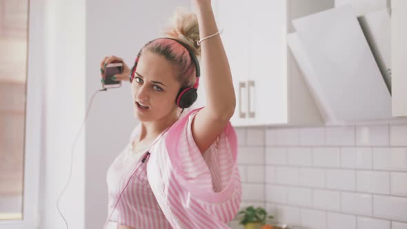 Happy Young Woman with Pink Hair Dancing in Kitchen Wearing Pink Pajamas and Listening to Music with alt