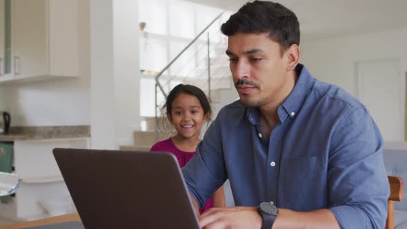 Happy hispanic father and daughter sitting at table looking at laptop alt