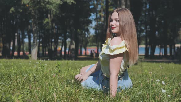 A Beautiful Girl Poses Sitting in the Park on the Grass alt