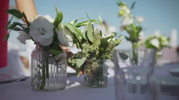 Female in Pink Shirt Putting Small Glass Jars with Flowers on Tbale with Plates and Cutlery alt