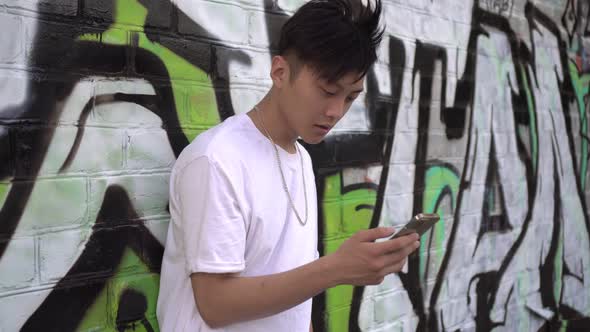 Young Asian Male Teenager Uses a Smartphone Against a Background of Street Graffiti alt