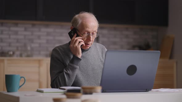 Old Man is Working with Laptop in Home Kitchen Calling By Phone for Consulting Medium Portrait of alt