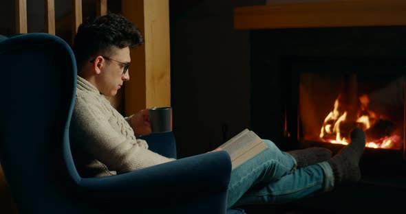 Man Reads Book and Drinks Tea Relaxing at Fireplace in Armchair at Winter Night