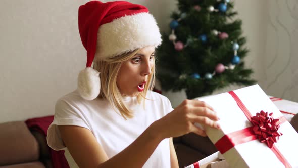 Shoked adorable woman in Santa Claus hat opening her Christmas present.