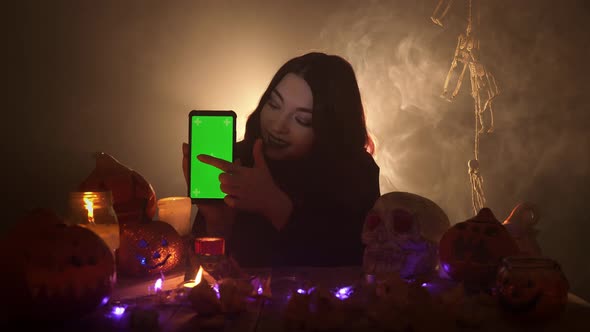 Woman Advertises a Mobile with Green Screen and Taps on It Surrounded By Halloween Decorations alt