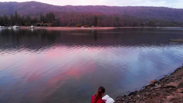 Aerial shot of a girl reading on a rock on a lake at sunset. alt