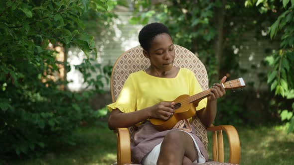 Portrait of Talented Relaxed African American Woman Playing Ukulele in Slow Motion Sitting on alt